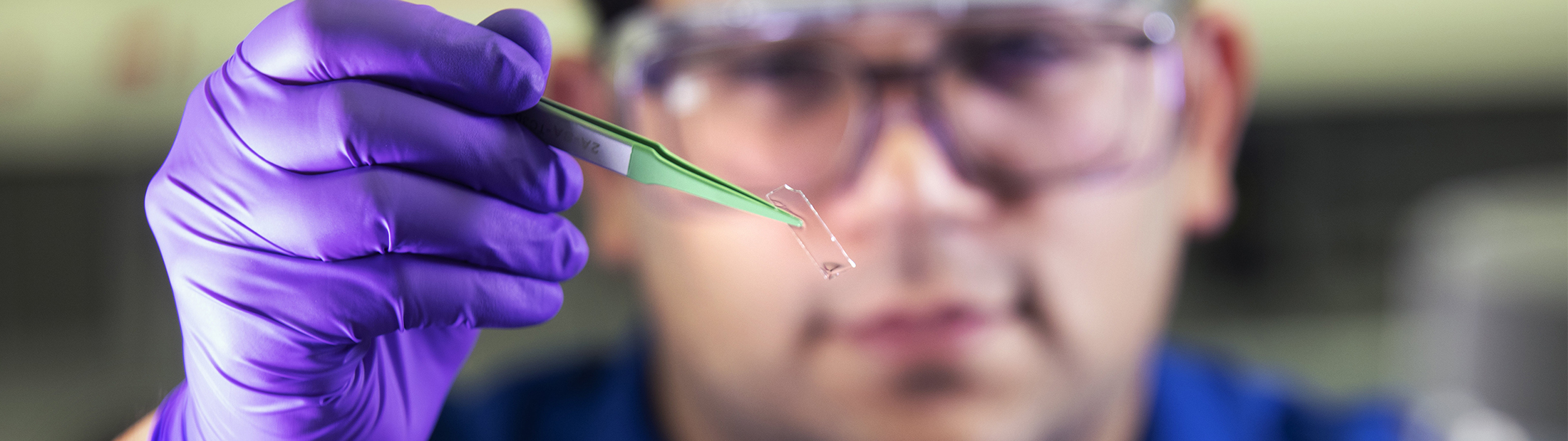 A person wearing safety glasses uses their gloved hand to hold up a small, transparent rectangle with a pair of tweezers.